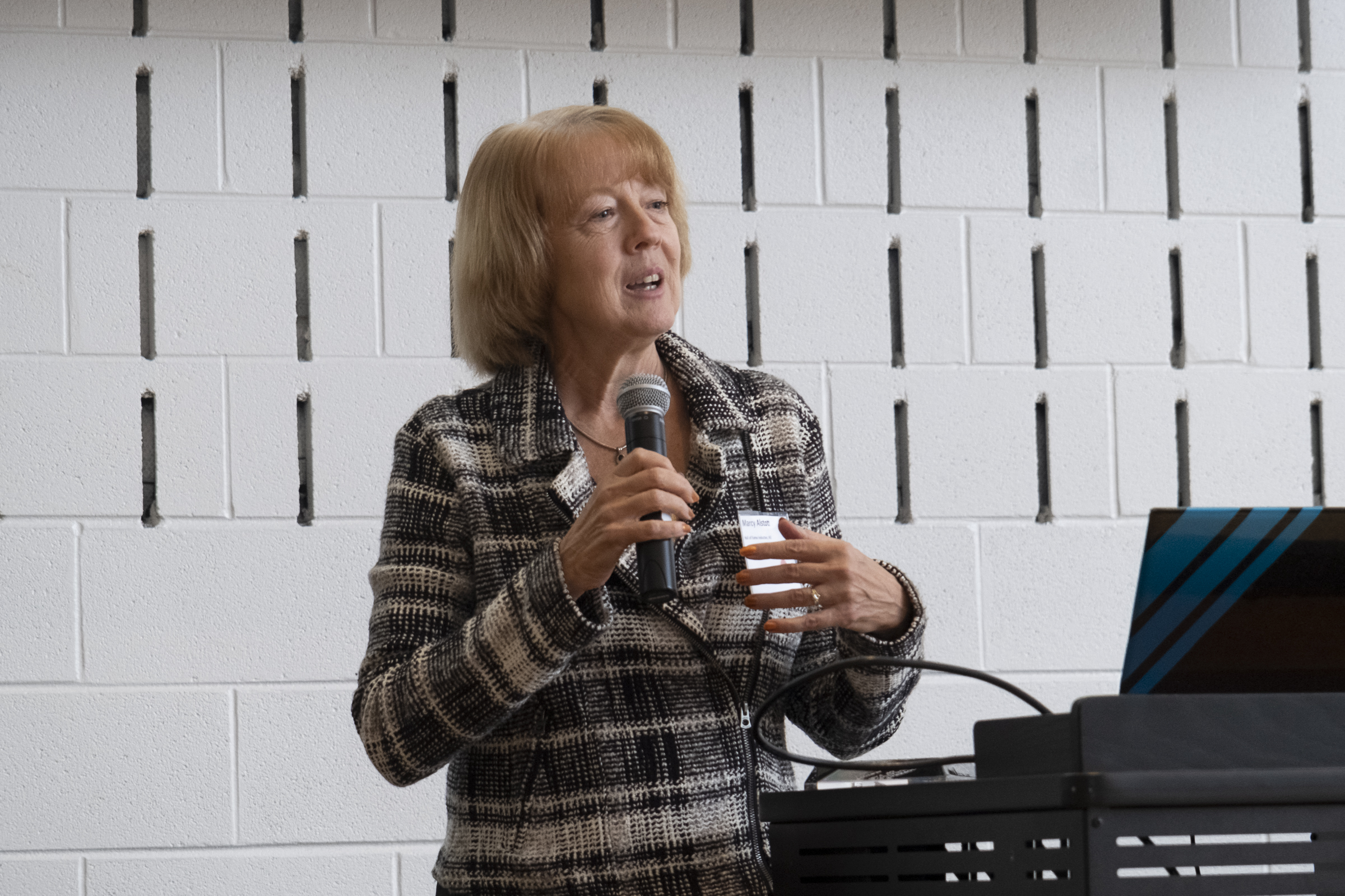 Marcy Alstott, supply chain and operations executive, speaking at an event wearing a checkered blazer standing by a black podium next to a white wall. 